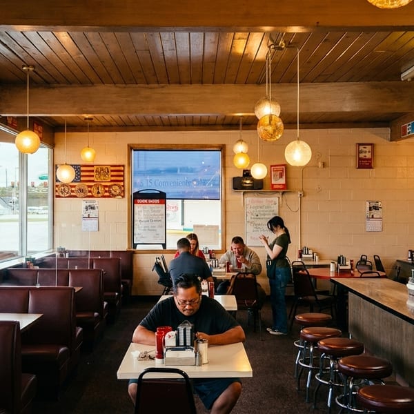 Diners eat in a retro-style restaurant with booths, a counter with stools, and large windows.